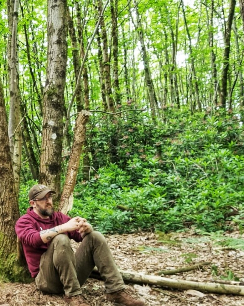 Sam sitting against a tree in dappled woodland, looking out, at rest.