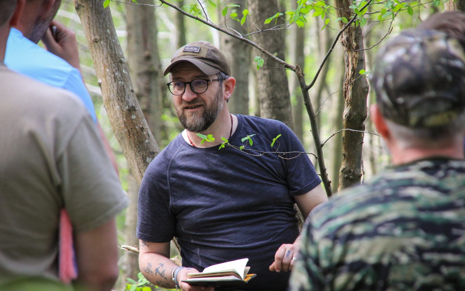 Sam sitting in the woods in conversation with a small group, a notebook in his hand.