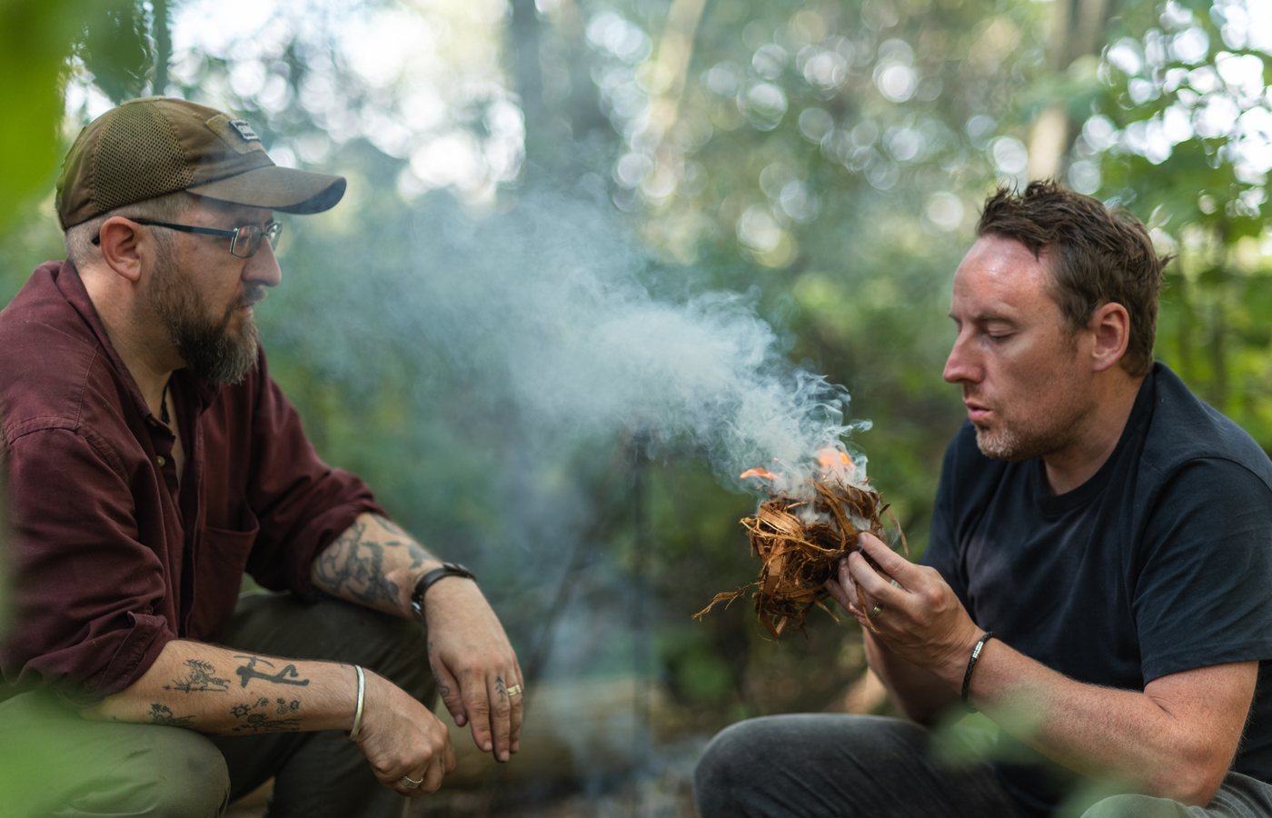 Sam looking on in the woods as a learner coaxes a smoking tinder bundle towards flame.